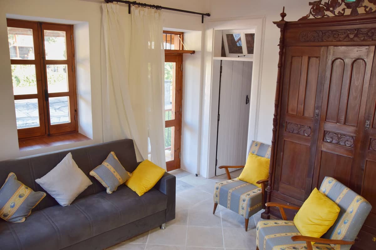 Una sala de estar con un sofá gris, dos sillones con cojines amarillos, una puerta de madera y una ventana con cortinas blancas.