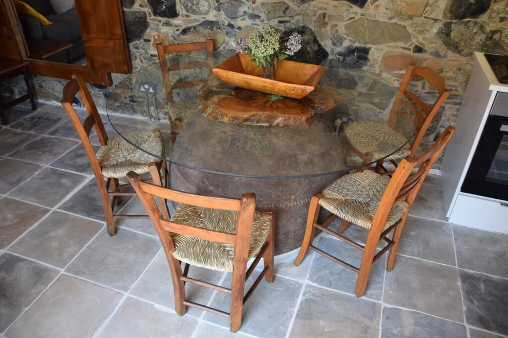 Una mesa redonda de madera rodeada de cuatro sillas de mimbre, con un muro de piedra de fondo.
