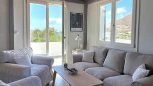 Ein Wohnzimmer mit einem gestreiften Sofa, einem beigen Sofa und einem Holztisch. Zwei Fenster bieten einen Blick ins Freie.