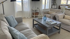 Ein modernes Wohnzimmer mit einer Couch, einem Couchtisch mit Büchern und einer Vase, einem Fernseher an der Wand und Blick ins Freie durch große Fenster.