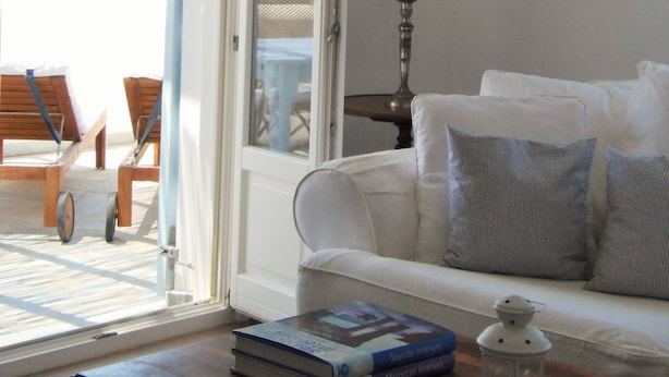 A room with a white sofa, a table with books, a lamp, and a view of the beach through an open door. - 'paradise' by Cnn - Delfini Bay - 3bed House (Syros)