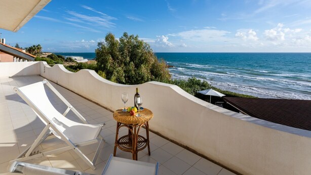 Un balcon avec un fauteuil de détente blanc, une table en osier avec une bouteille et des verres, et une vue sur l’océan.