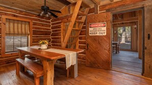 L’intérieur d’une cabane en rondins, avec une table en bois, des bancs, un ventilateur de plafond et un panneau d’avertissement concernant la présence d’une mine non explosée.