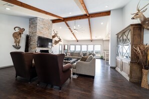 Ein großzügiges Wohnzimmer mit dunklem Holzboden, einem Steinkamin und einem großen Fernseher an der Wand. Zwei braune Ledersessel und ein beiges Sofa sind ebenfalls vorhanden.