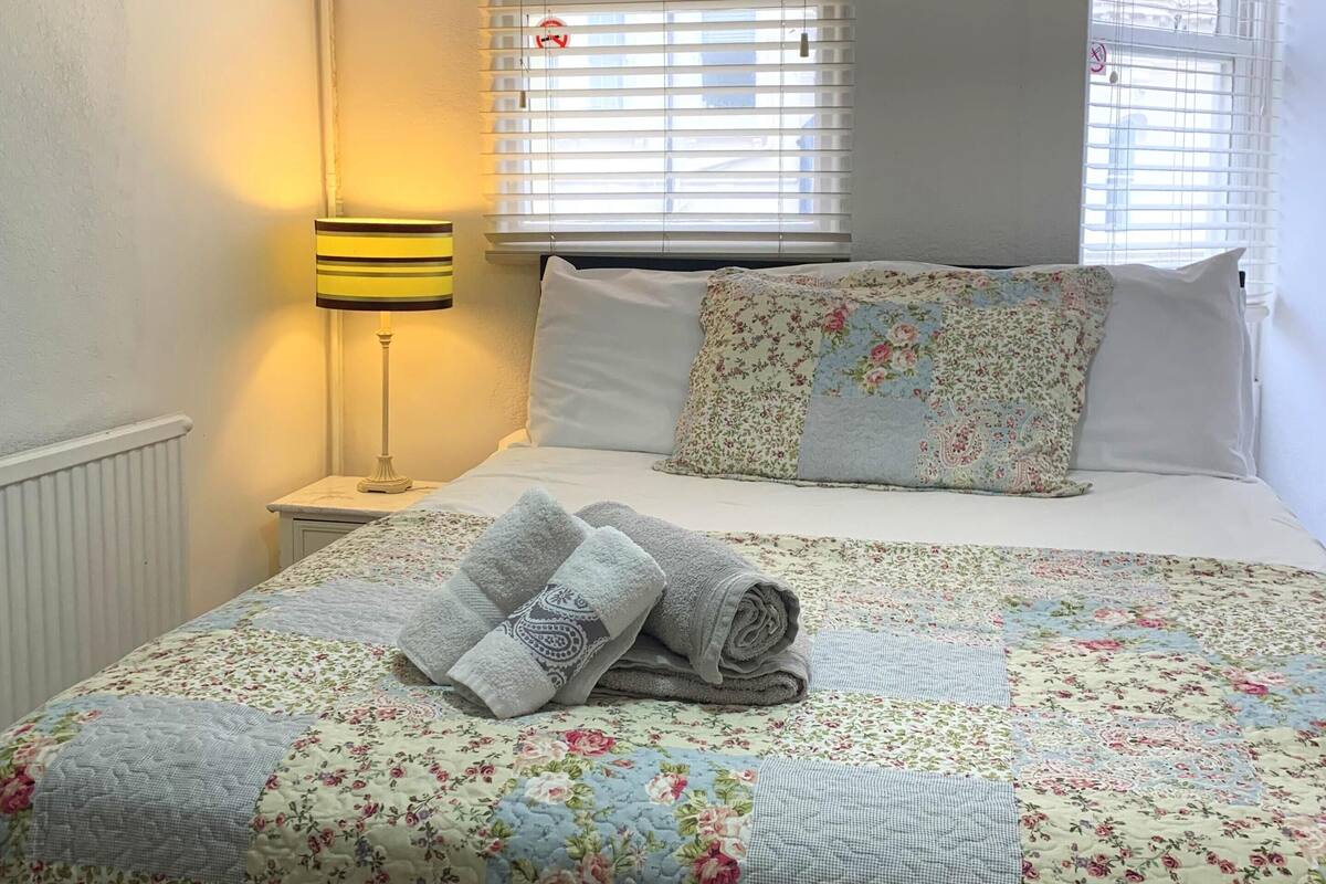 A neatly made bed with a floral duvet, a bedside lamp, and a window with blinds.