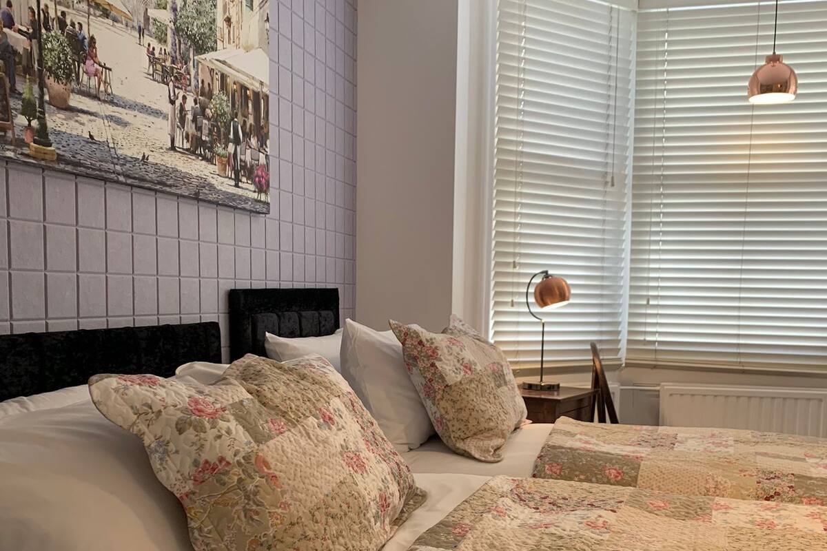A bedroom with a large bed, floral bedding, a cityscape painting on the wall, and a window with blinds.