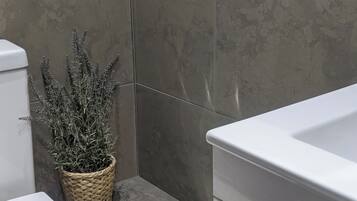 Un baño con inodoro y lavamanos blancos, un bote de basura y una planta en maceta.