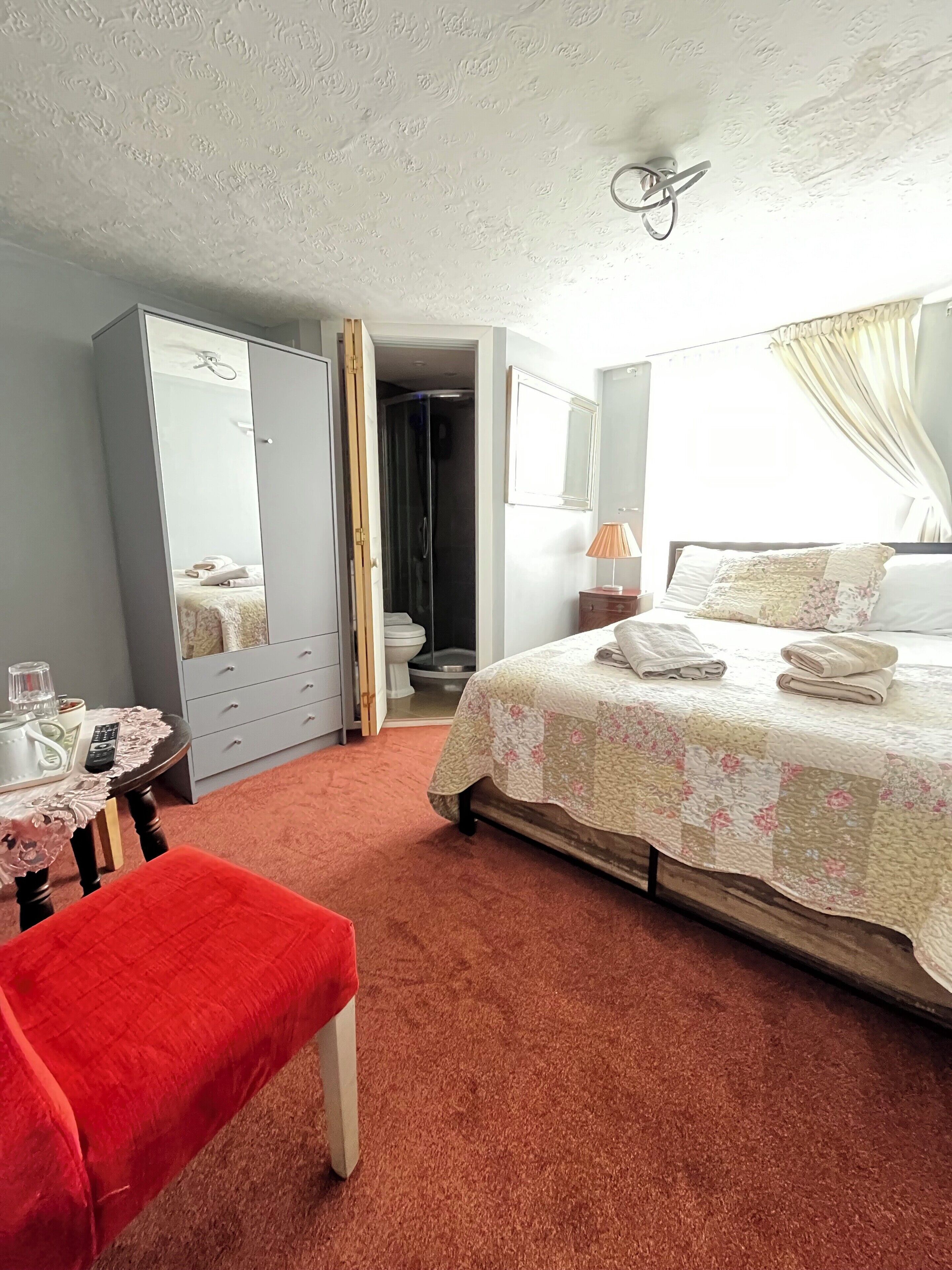 Ein Schlafzimmer mit einem Bett, einem roten Sessel, einem Kleiderschrank, einem Spiegel, einem Nachttisch und einem durchgehenden Türbogen sichtbaren Badezimmer.