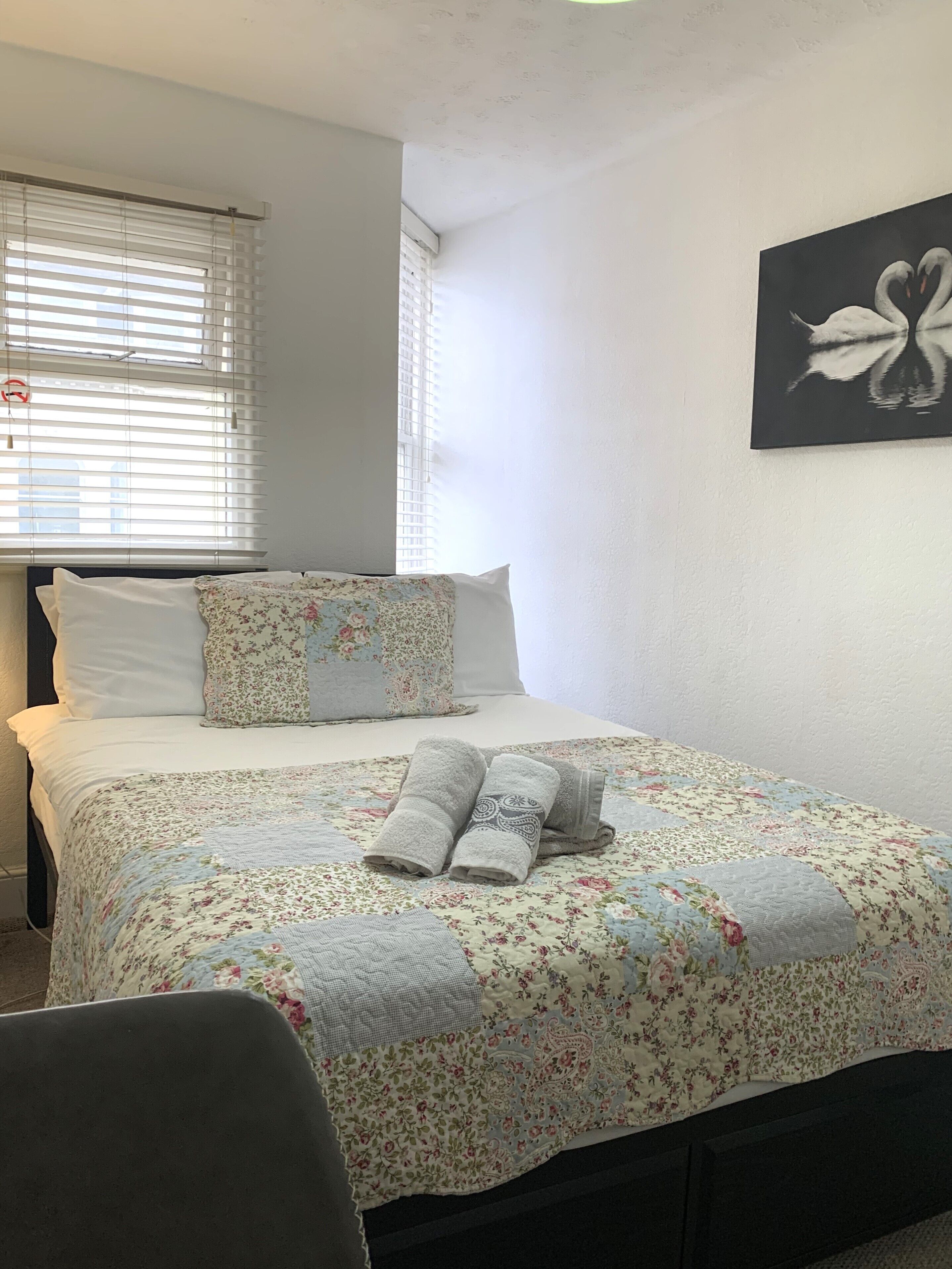 A neatly made bed with floral bedding and rolled towels, a wall art of swans, and a window with blinds.