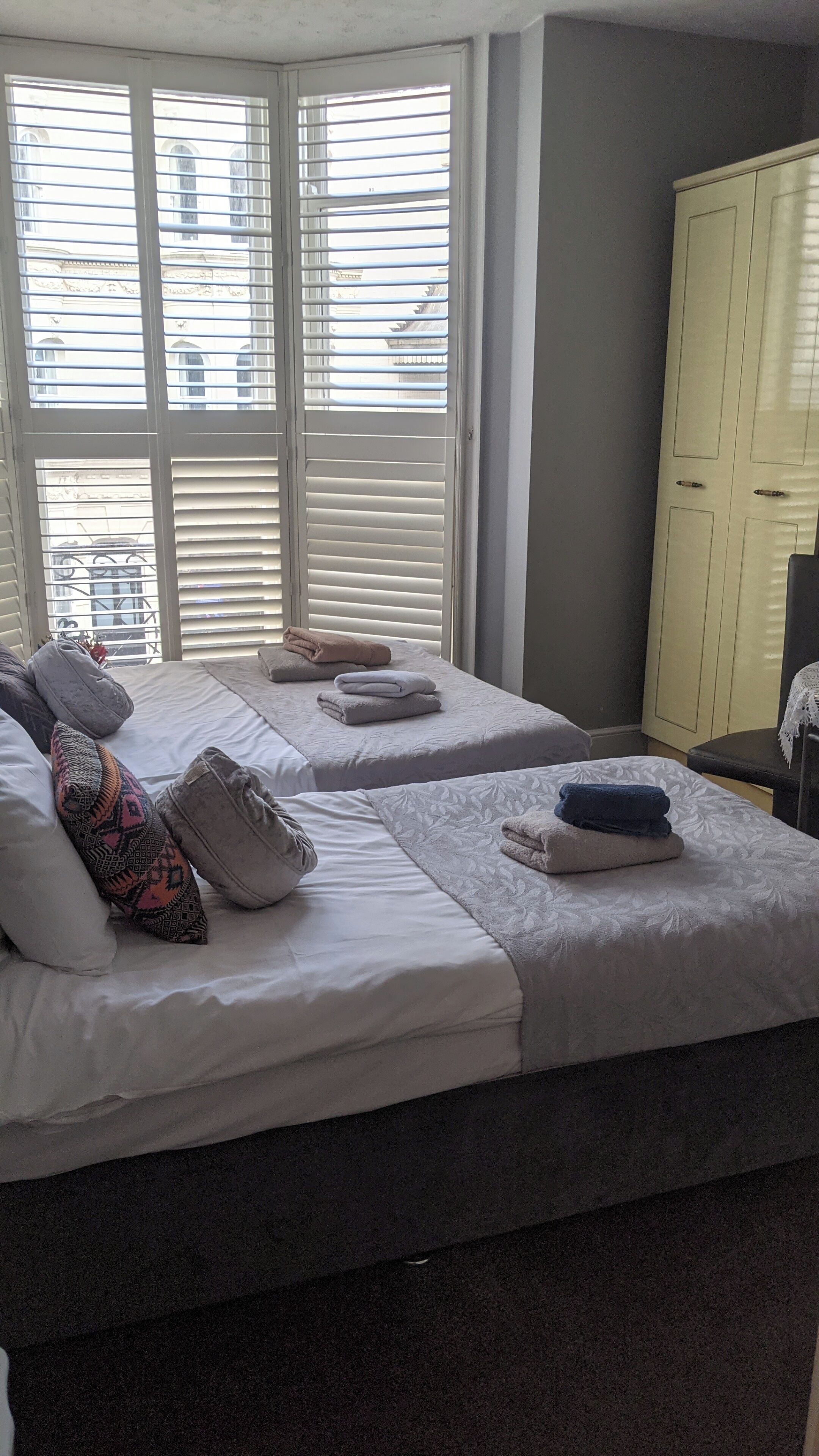Quarto de hotel com duas camas, uma janela grande com persianas, um guarda-roupa e vista para edifícios.