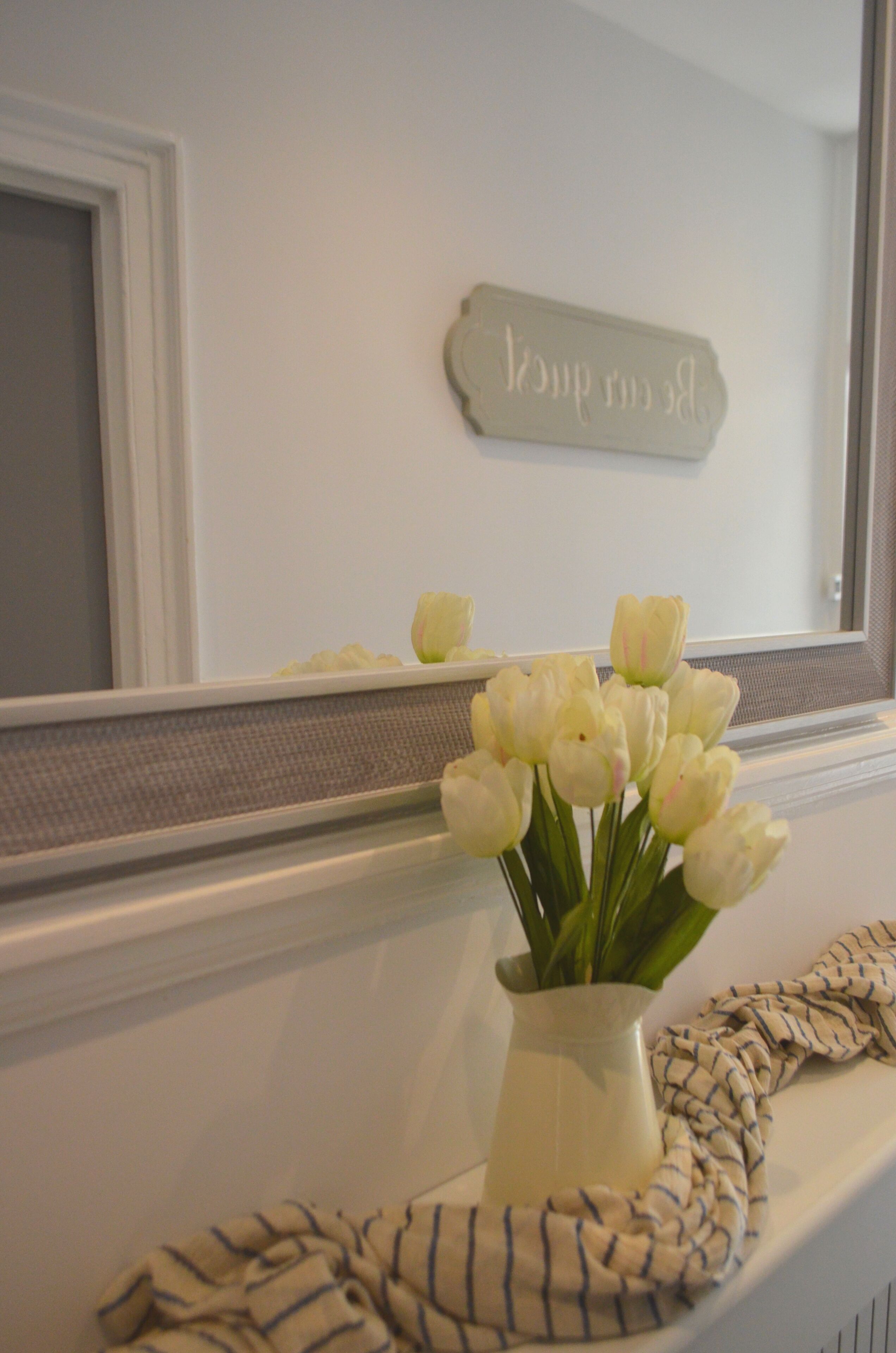 A bathroom with a white vanity, a vase of white tulips, and a mirror with a sign above it.