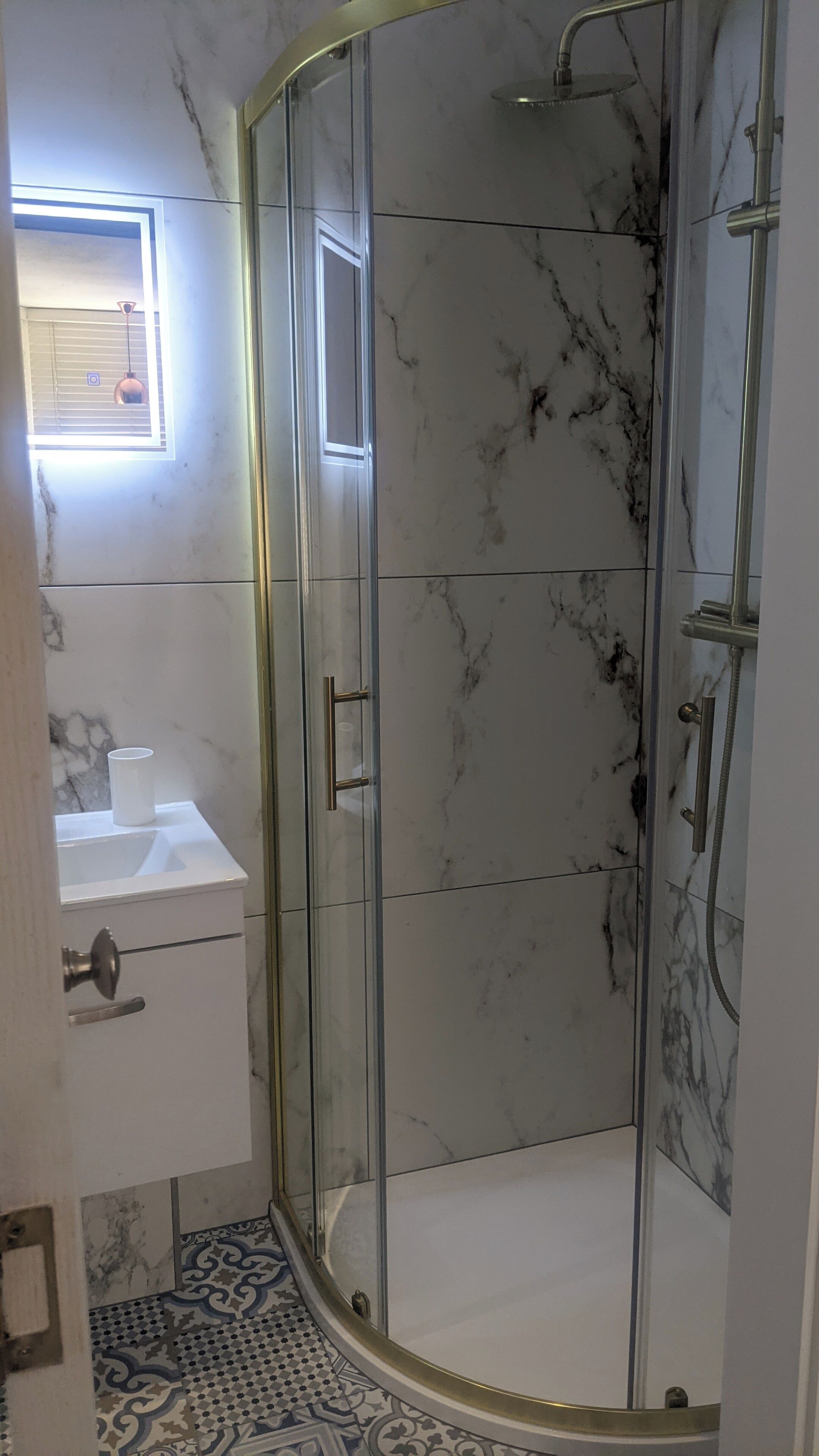 Un baño con ducha acristalada, lavabo blanco y suelo estampado.