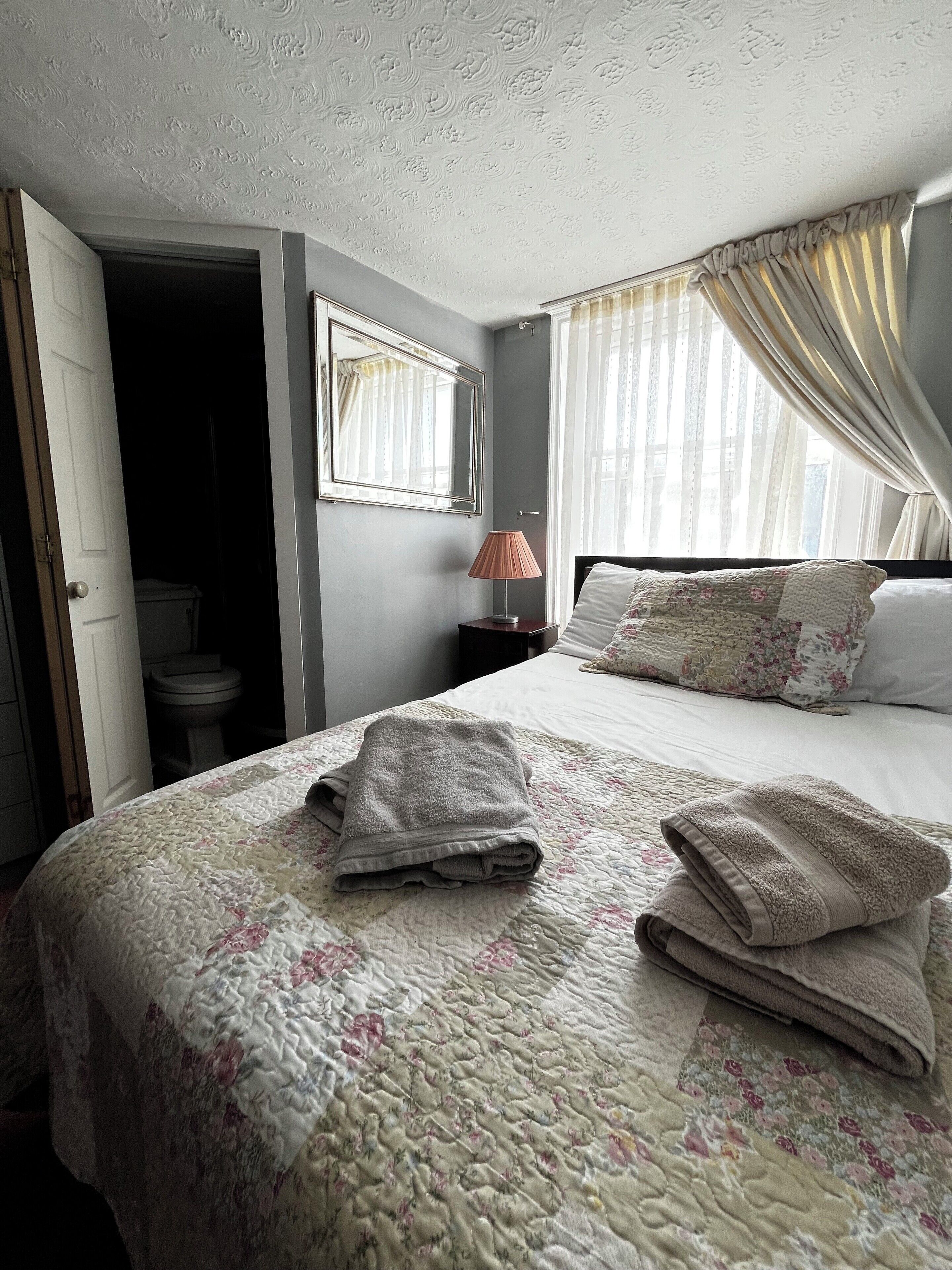 A hotel room with a bed, a nightstand, a lamp, a window with curtains, and a door leading to a bathroom.