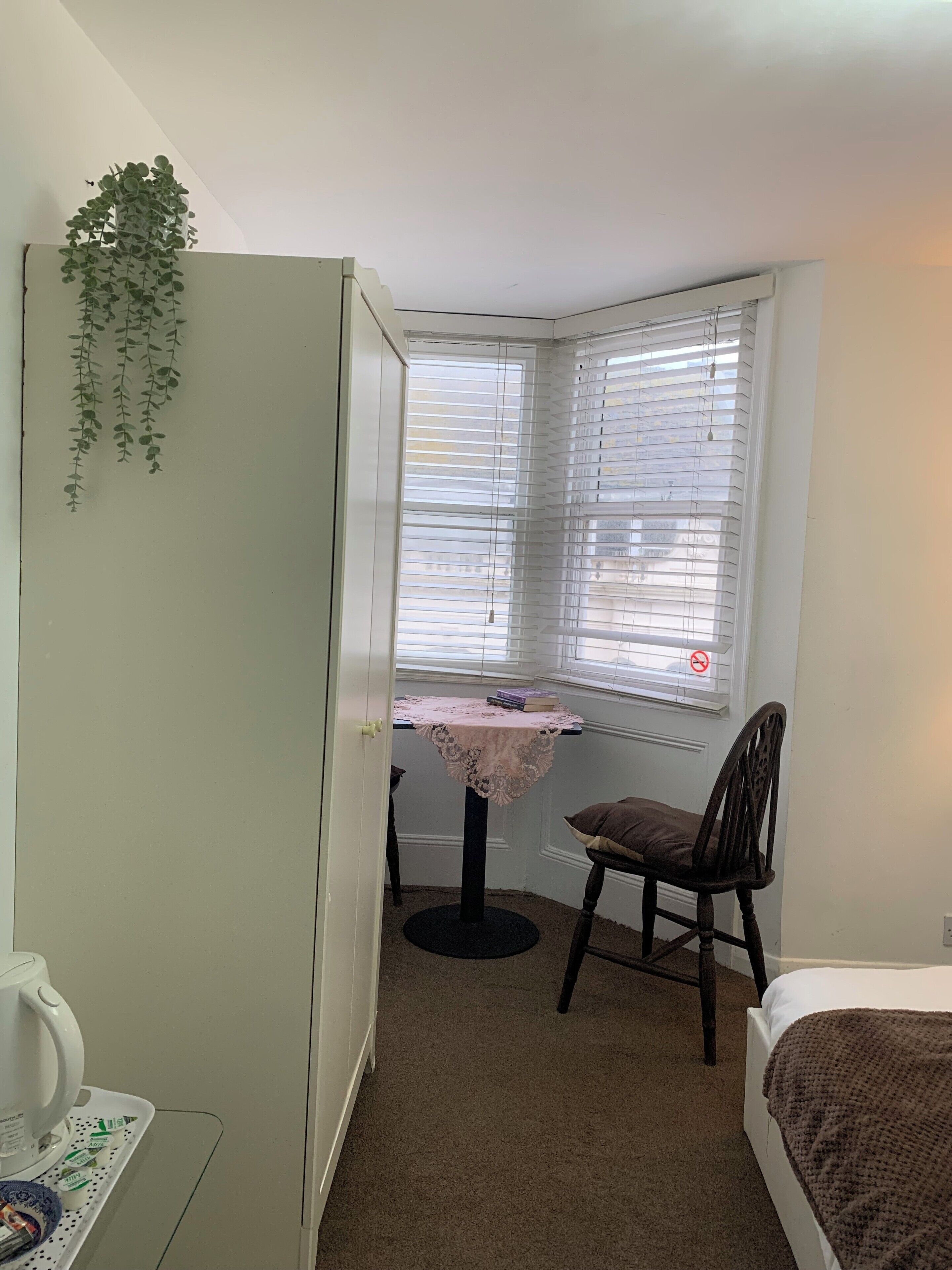 A small, neatly arranged bedroom with a single bed, a table with a lace tablecloth, a chair, and a window with blinds.