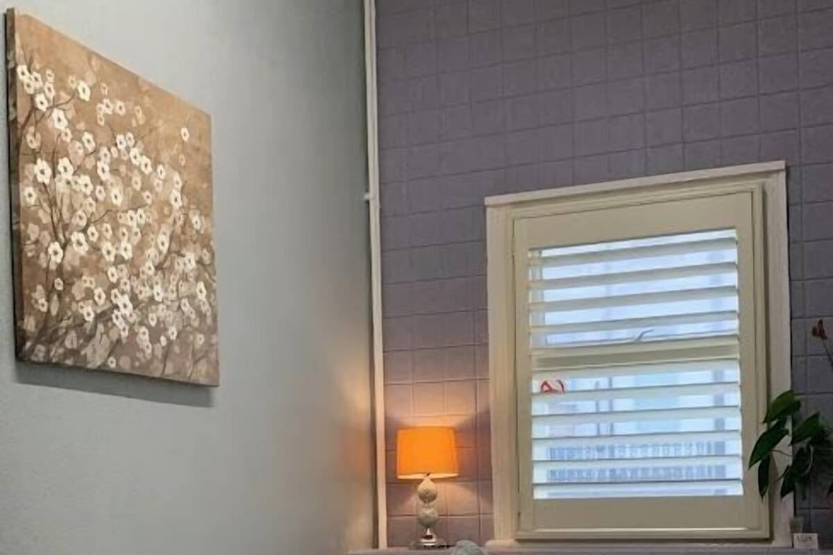 A bedroom with a bed, a nightstand, a lamp, a window with blinds, and a wall painting.