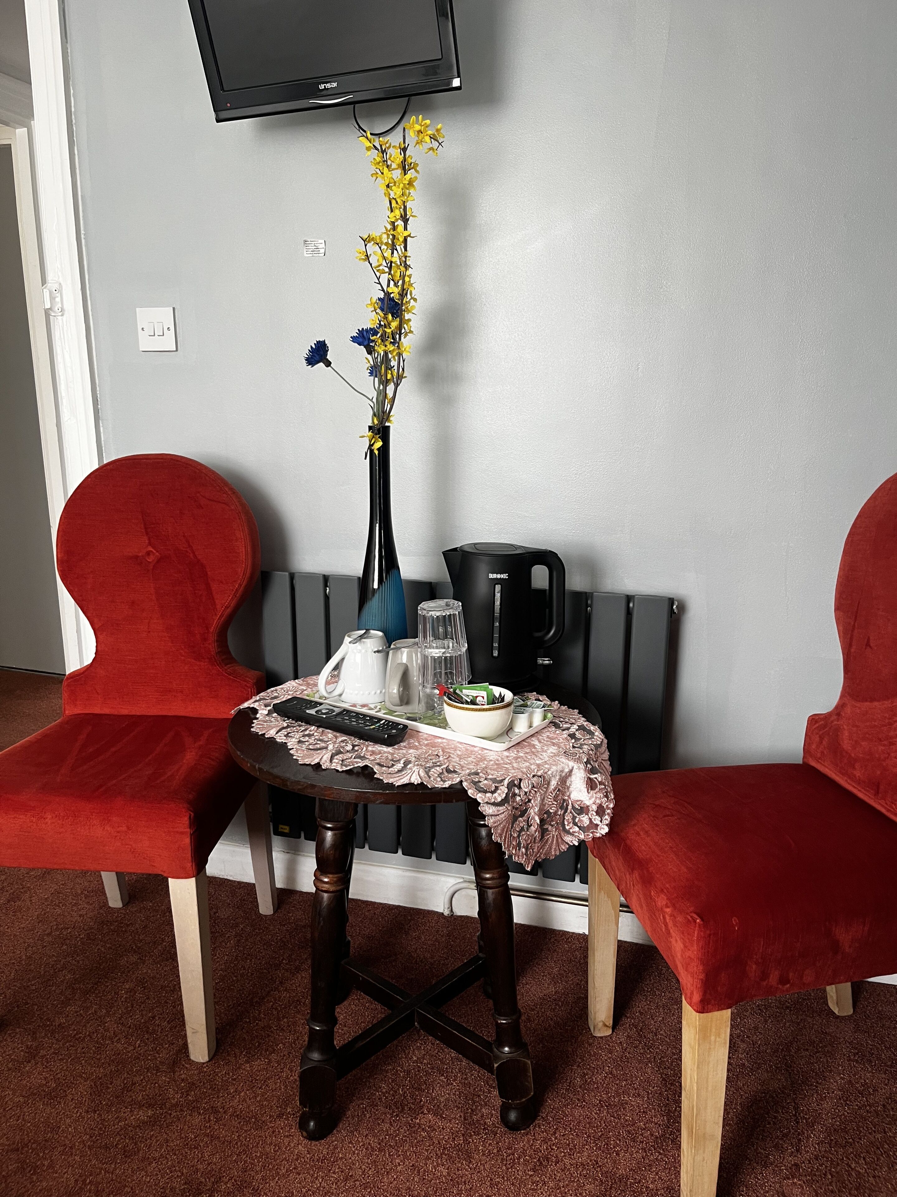 A small room with a table set for two, a vase with yellow flowers, a kettle, and a remote control.