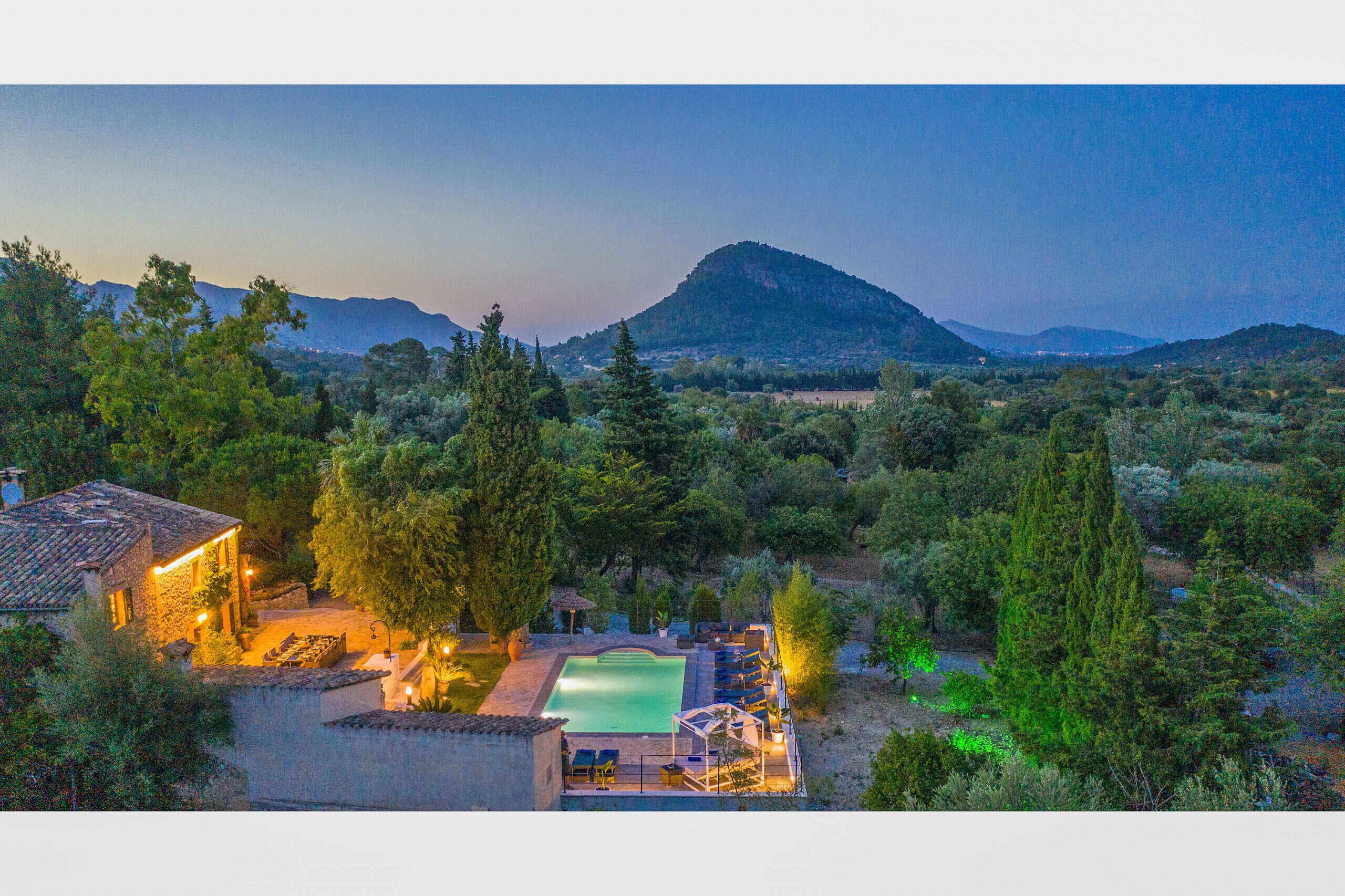 A pool area with lounge chairs, a small building with lights, and a mountainous backdrop.