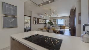 A modern kitchen with a built-in stove, a dining area with a wooden table and chairs, and a view of the ocean through large windows.
