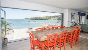 A dining area with a large table and orange chairs, overlooking a bay with boats and a hilly landscape. - Ynys Faelog (Bangor)