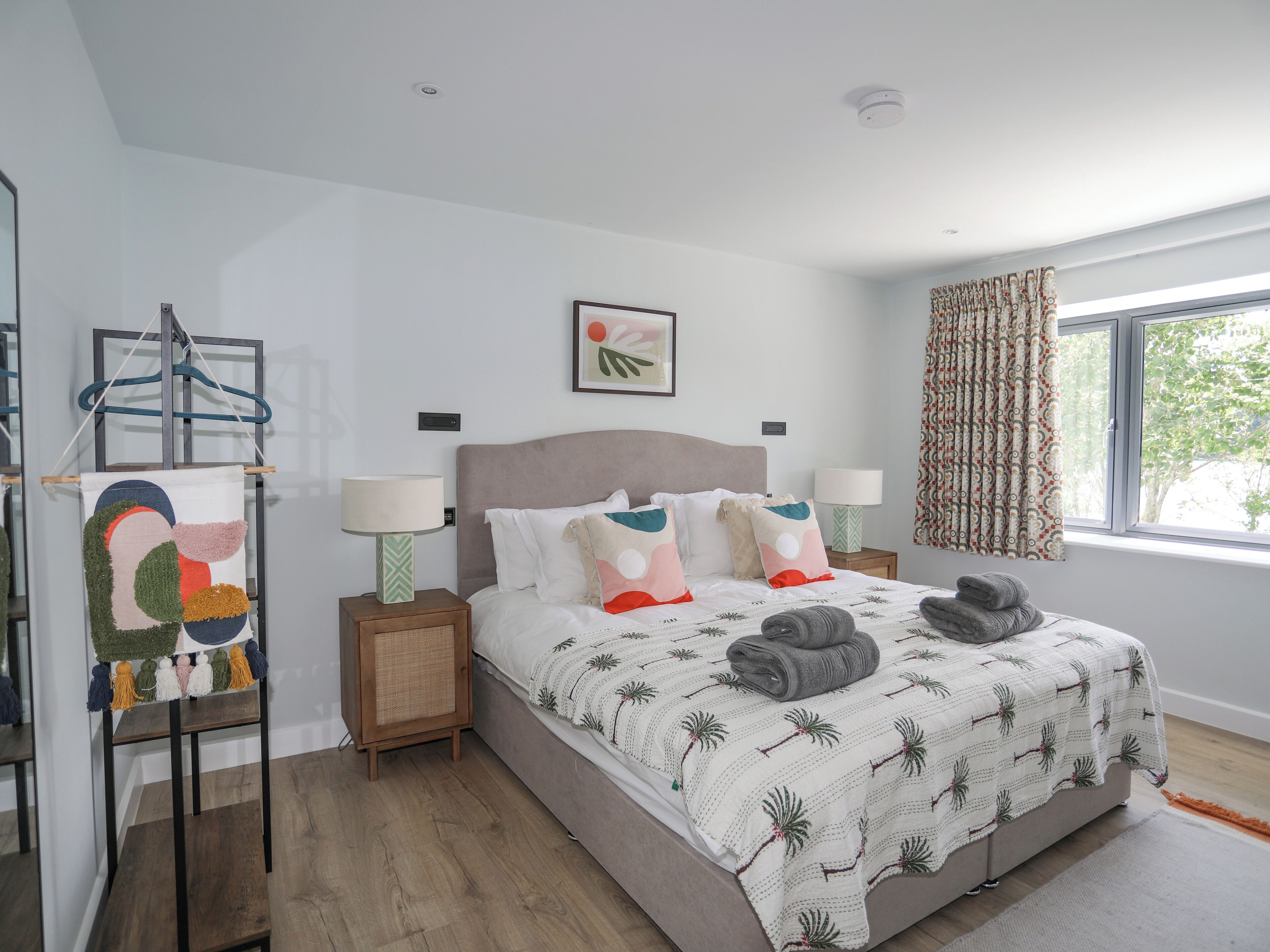 Une chambre moderne avec un grand lit, deux tables de chevet, une lampe fixée au mur, une fenêtre avec des rideaux et une œuvre d’art abstraite suspendue au mur.