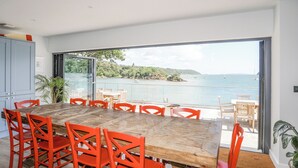 A dining area with a large wooden table and red chairs, overlooking a scenic bay with boats. - Ynys Faelog (Bangor)