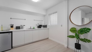 Una cocina moderna con armarios blancos, un microondas, una cafetera y un lavabo. Hay una planta pequeña y un espejo redondo en la pared.