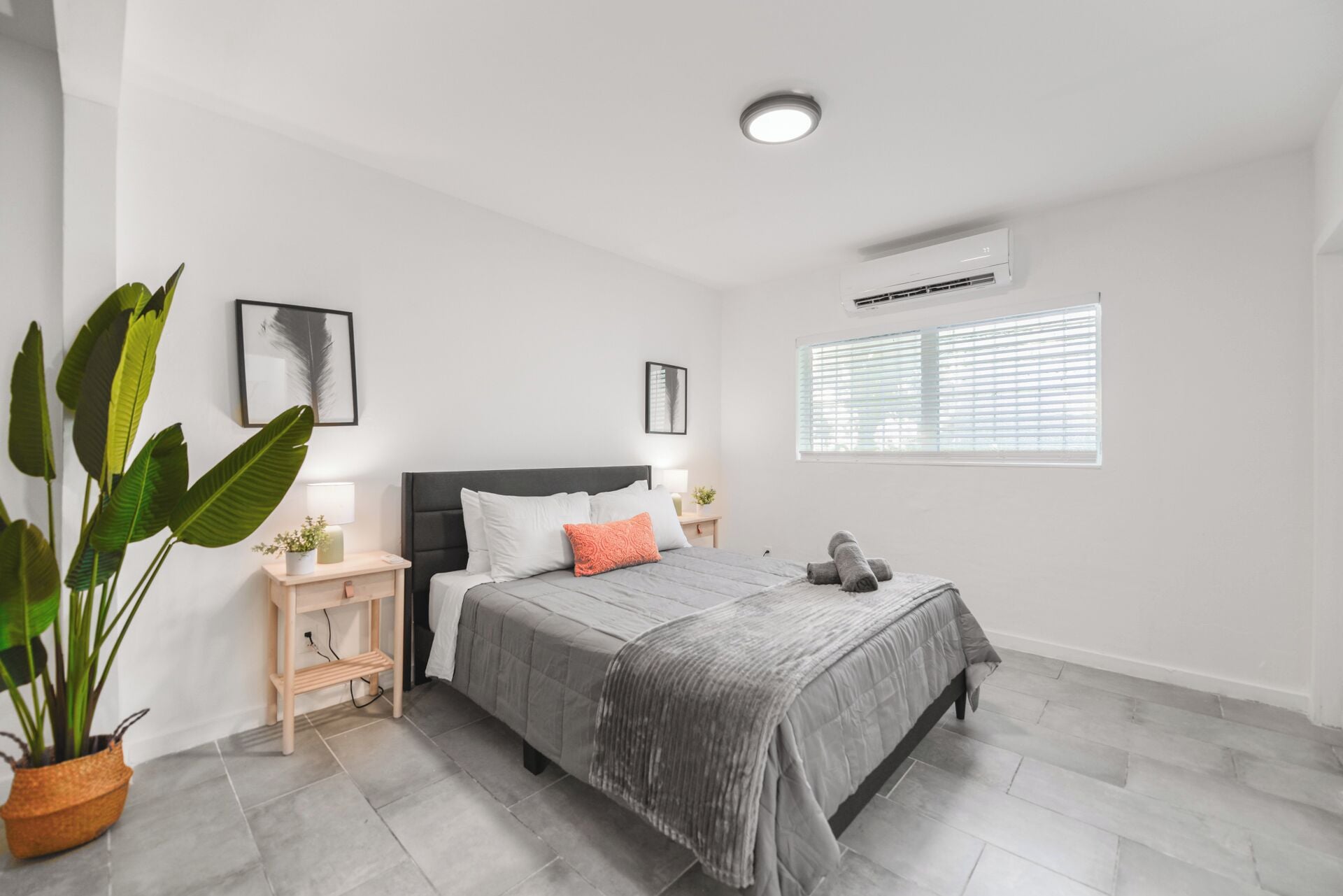 Un dormitorio moderno con una cama, mesitas de noche, una planta en maceta y una ventana con persianas.