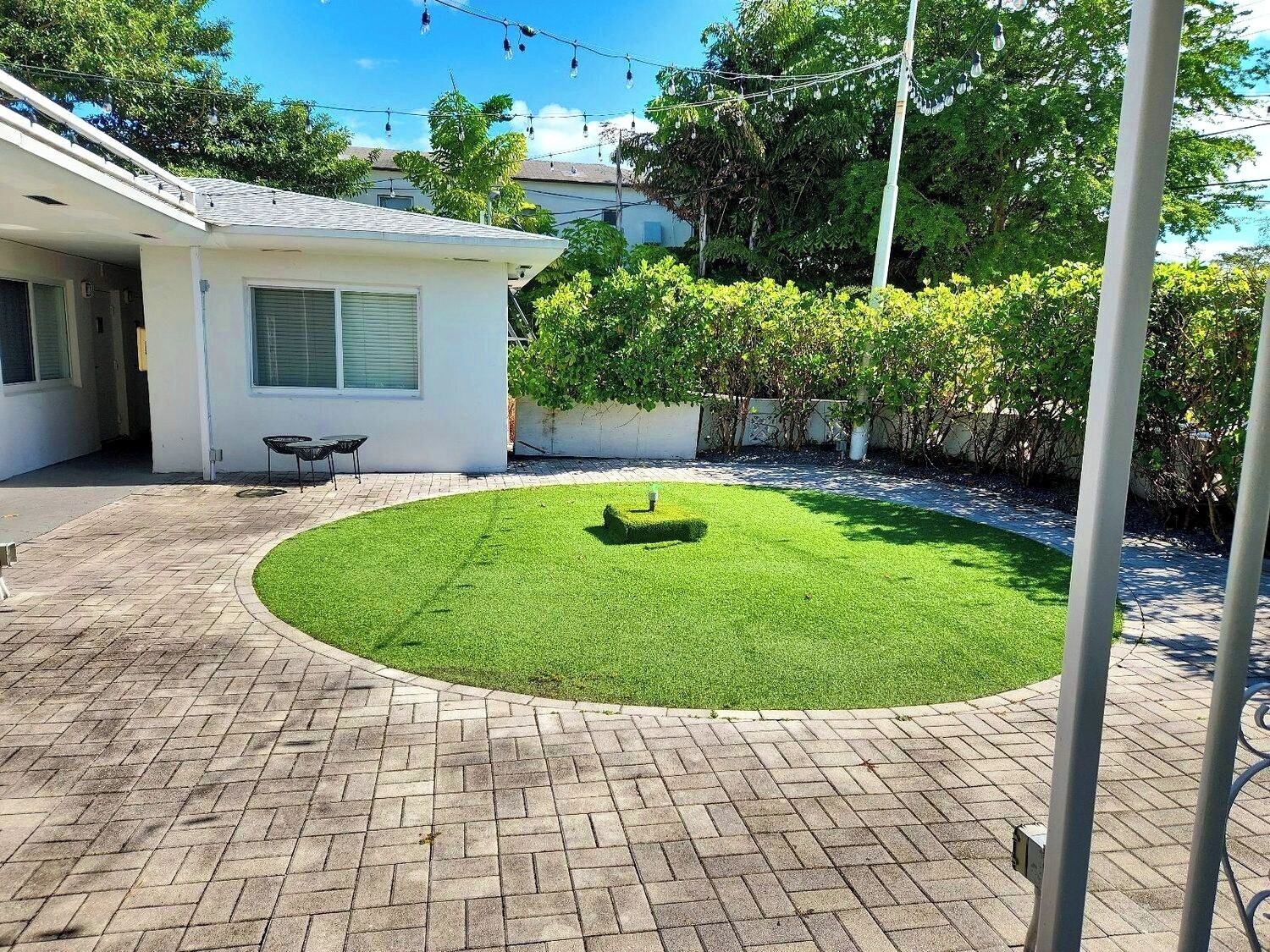 A well-maintained backyard with a paved patio, a small lawn, and a house with a white exterior and a window with shutters.