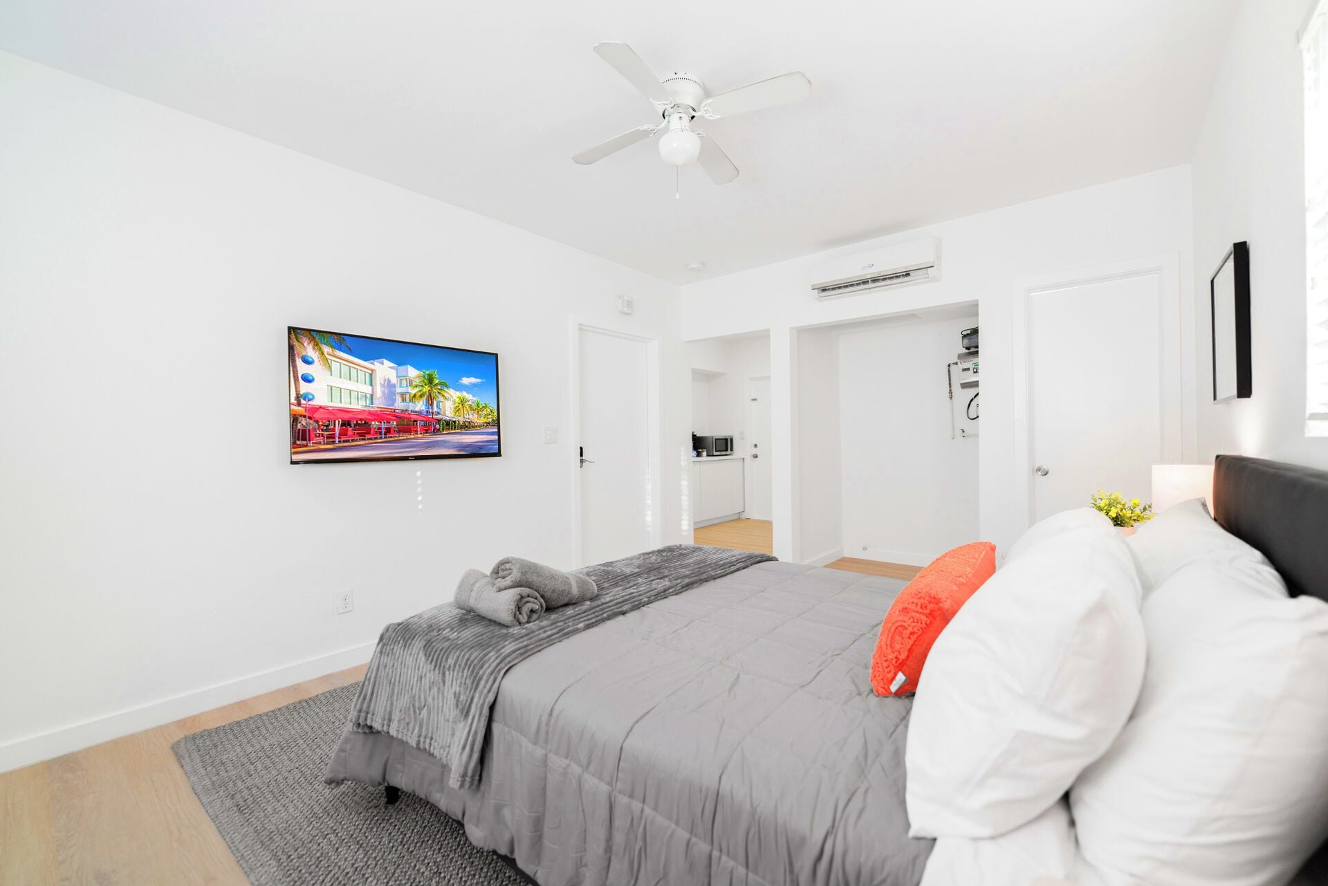 Un dormitorio moderno con cama, ventilador de techo, televisión y vista a una cocina y pasillo.