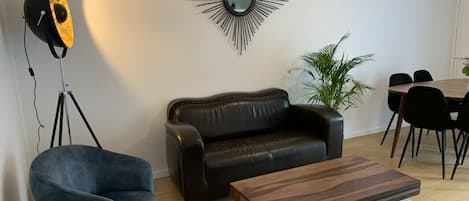 Una sala de estar moderna con un sofá de cuero, un sillón azul, una mesa de centro de madera, una lámpara de pie y un comedor con mesa y sillas.