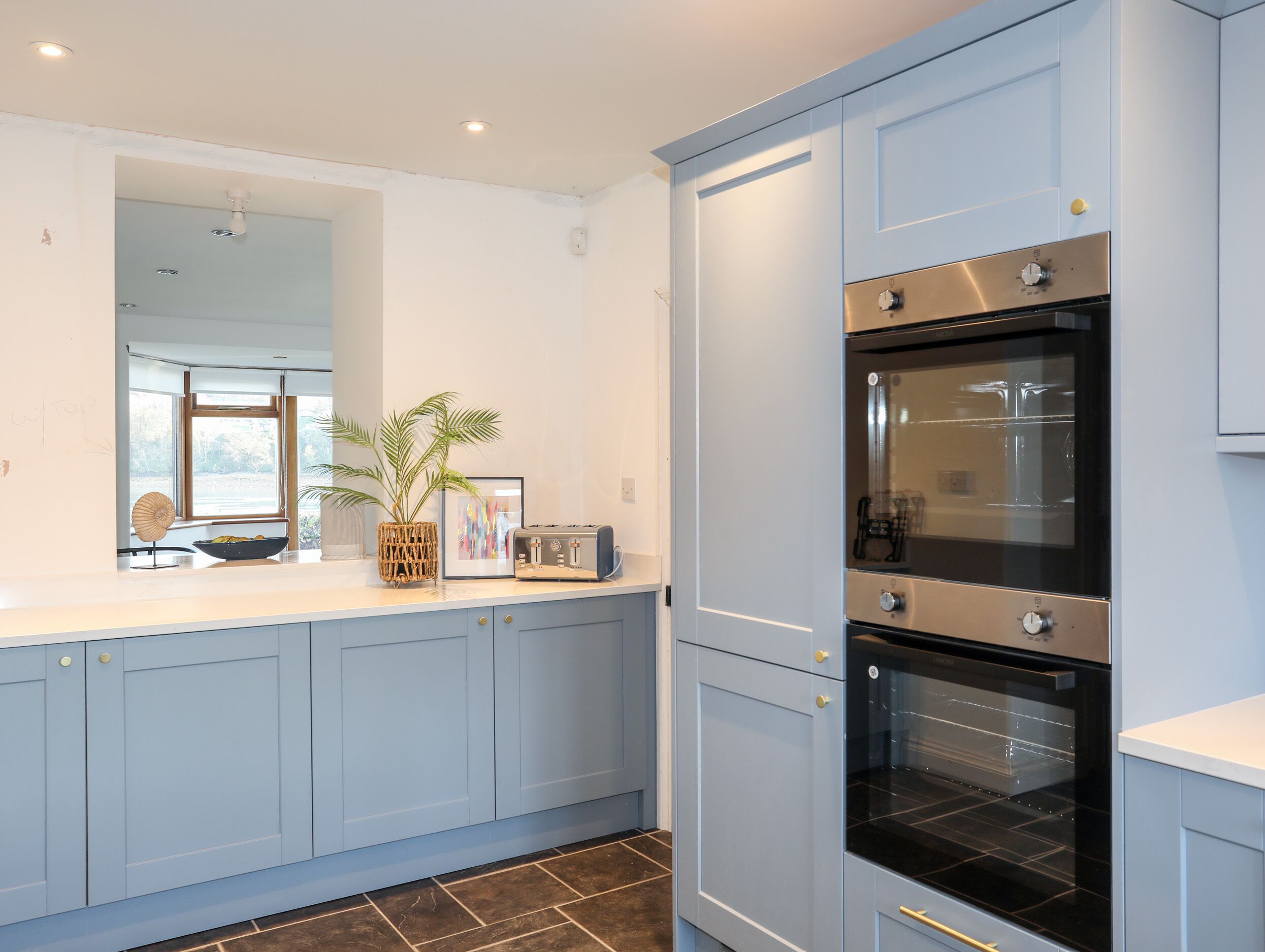 A modern kitchen with light blue cabinets, a double oven, and a large window providing natural light.