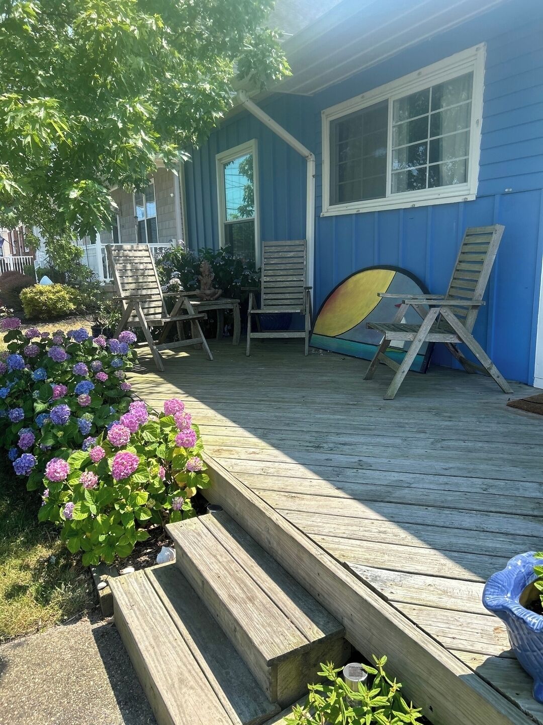 Una terraza de madera con una casa azul, una silla de madera, una tabla de surf y flores de hortensia.