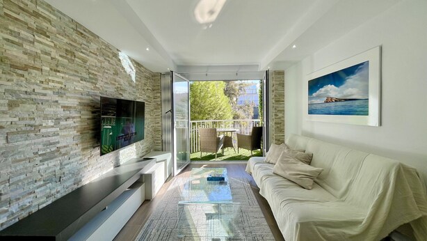 A modern living room with a stone wall, a flat-screen TV, a glass coffee table, a beige sofa, and a painting of a coastal scene. - DELUXE CENTER R146 (Benidorm)