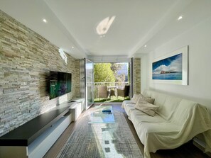 Un salon moderne avec un mur en pierre, un téléviseur  à écran plat, une table basse en verre, un canapé beige et un tableau représentant un paysage côtier.
