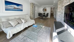 A modern living room with a white sofa, a glass coffee table, and a dining area with a stone accent wall. - DELUXE CENTER R146 (Benidorm)