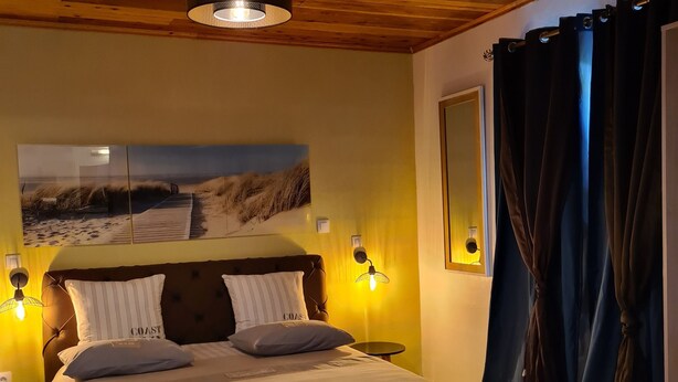 A bedroom with a wooden ceiling, a bed with white bedding, a painting of a beach scene, and a nightstand with a lamp. - Les Chambres et table d'hôte du Voyageur/ chambre citronnelle (La Plaine Des Palmistes)