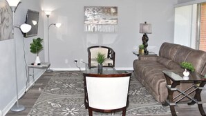 Un salon moderne avec un canapé en cuir marron, une table basse en verre, une table à manger avec des chaises et un tapis à motifs.