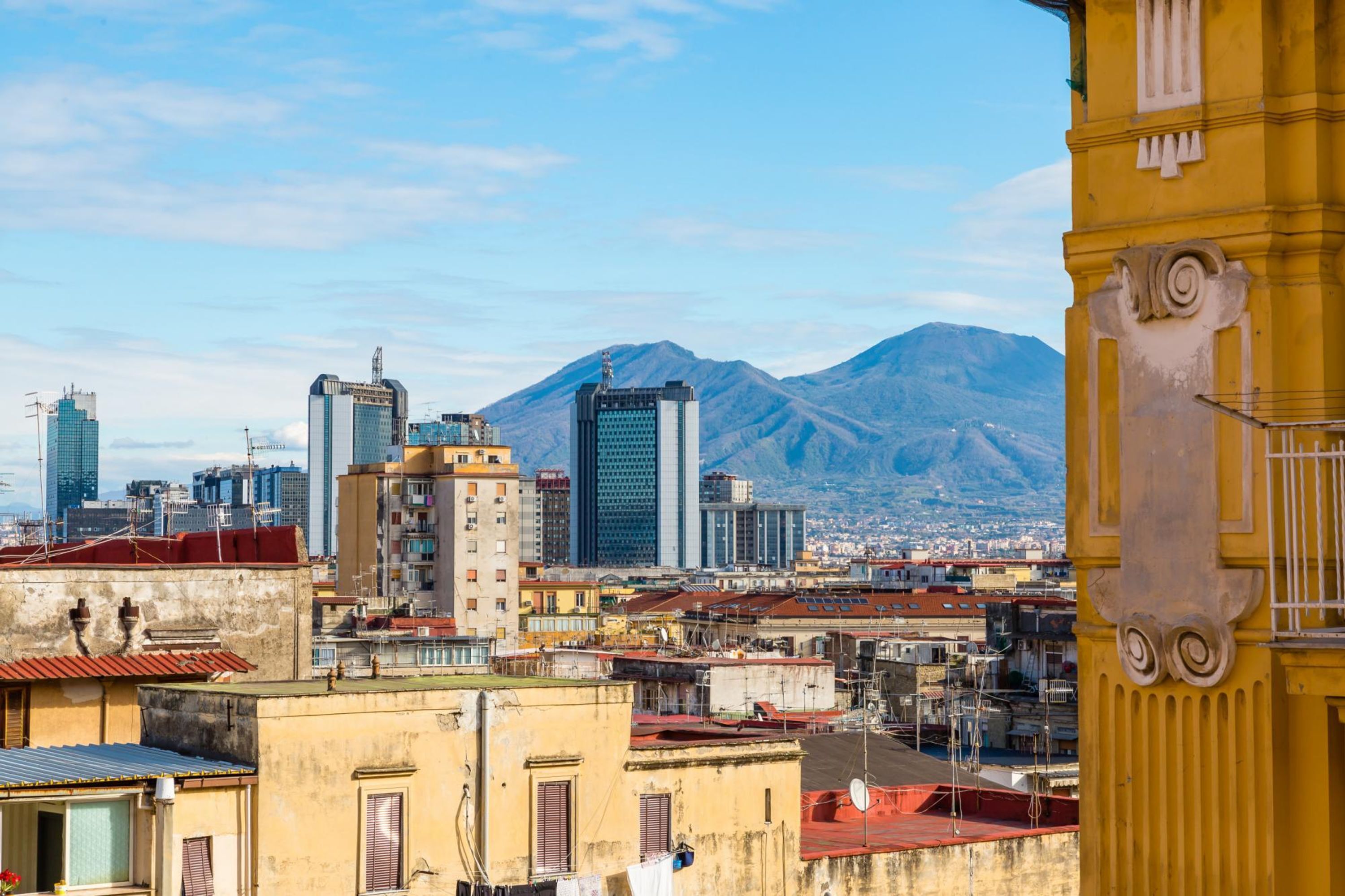 Un paisaje urbano con una montaña al fondo, que muestra varios edificios, incluyendo un edificio amarillo destacado con detalles ornamentales.
