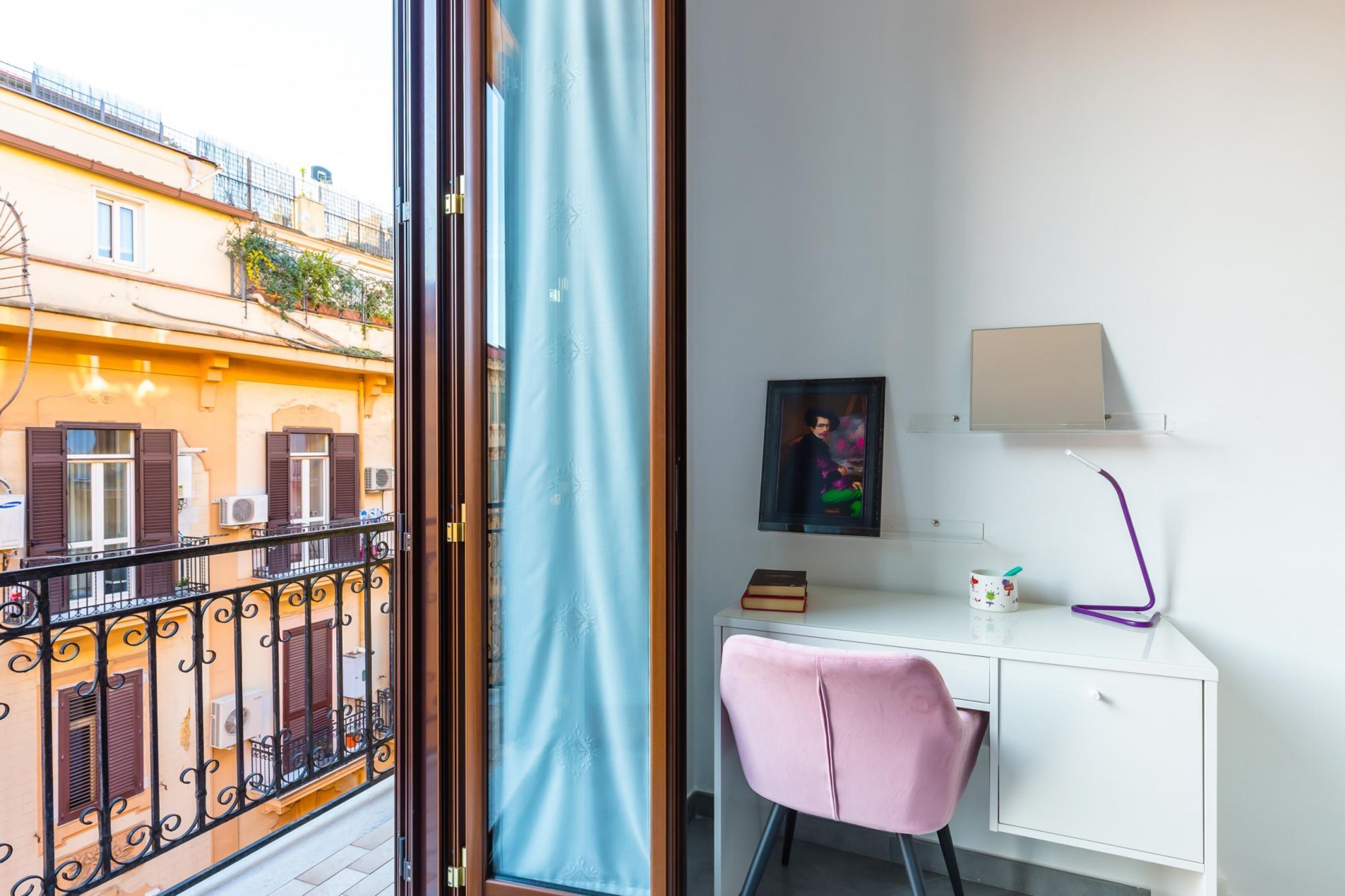 A room with a balcony, a desk with a chair, a framed picture, a lamp, and a small shelf with items.