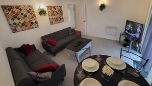 A living room with a sofa, a coffee table, and a dining area set with plates and glasses. There are two wall-mounted paintings and a TV on a stand. - Captivating 2-bed Apartment in Southend-on-sea (Southend-on-Sea)