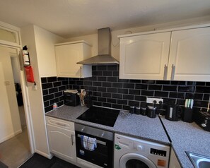 A compact kitchen with white cabinets, a black tiled backsplash, a built-in oven, a washing machine, and a stainless steel range hood. - Captivating 2-bed Apartment in Southend-on-sea (Southend-on-Sea)