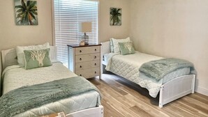 Une chambre avec deux lits, une commode, un ventilateur de plafond et des œuvres d’art représentant des palmiers sur les murs.