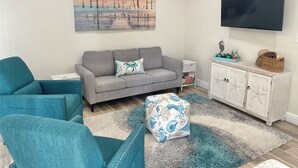 Un salon avec un canapé gris, des fauteuils turquoise, un meuble blanc décoré d’étoiles de mer et un ventilateur de plafond.