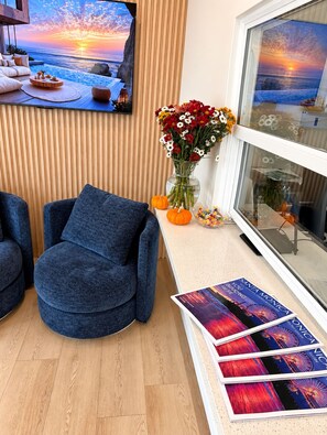 A modern living room with a large window, a blue armchair, a wooden floor, a coffee table with a vase of flowers, and a framed picture on the wall. - Rest Haven Motel (Santa Monica)