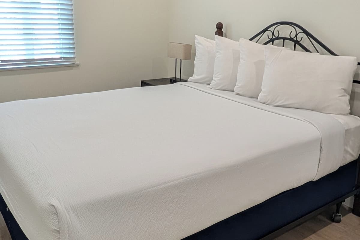 Un dormitorio con cama, mesita de noche, lámpara y ventana con persianas.