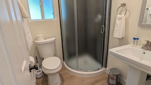 A bathroom with a toilet, a glass shower enclosure, a sink, and a toilet paper holder. - Rest Haven Motel (Santa Monica)
