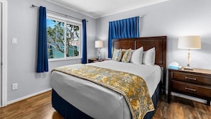 A hotel room with a large bed, a wooden nightstand with a lamp, a window with curtains, and a view of greenery outside. - Rest Haven Motel (Santa Monica)