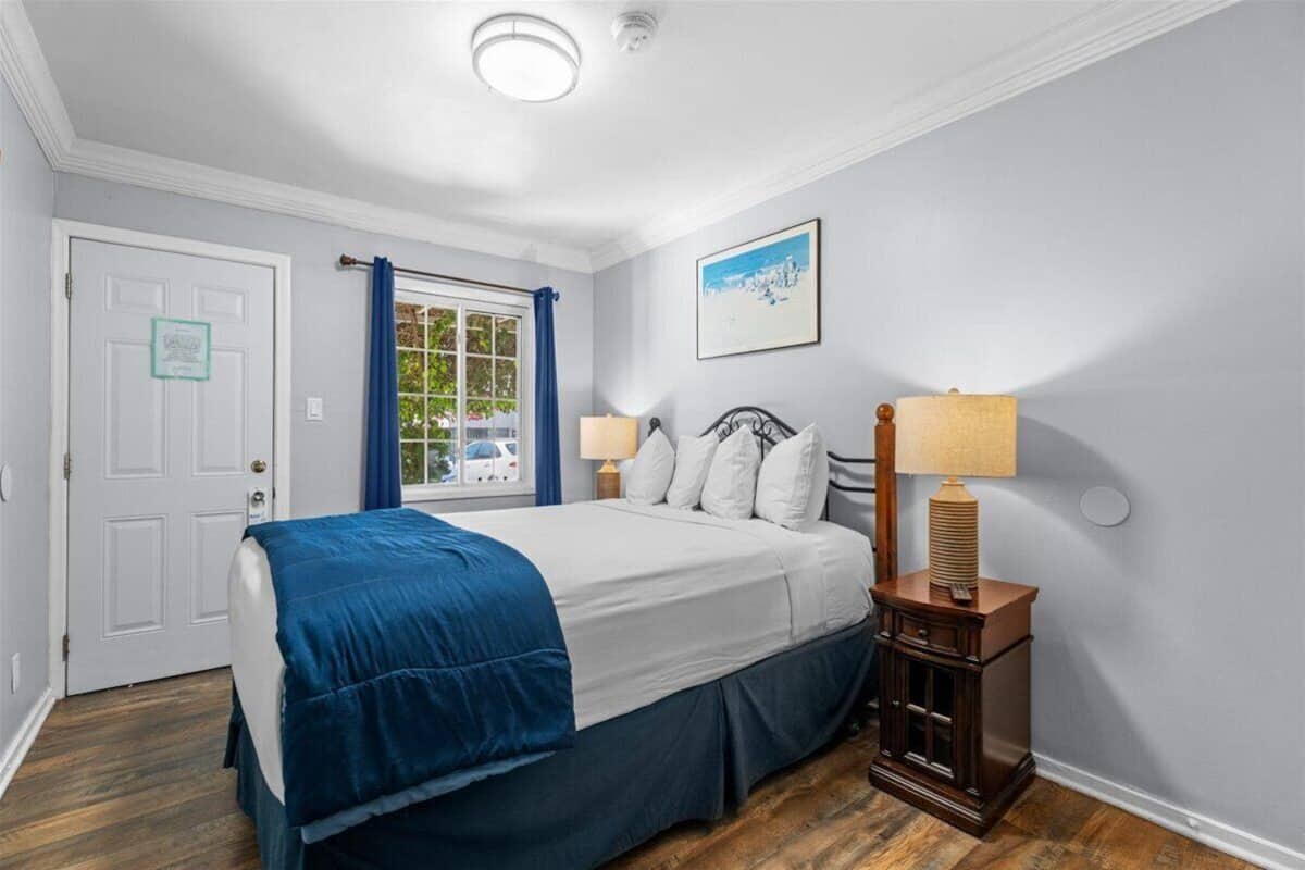 Un dormitorio con cama, mesita de noche, lámpara, ventana con cortinas, puerta y un cuadro en la pared.