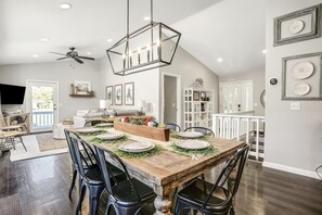Un comedor con una mesa de madera dispuesta para seis personas, rodeada de sillas negras. Se observa un ventilador de techo y lámparas colgantes sobre la mesa.