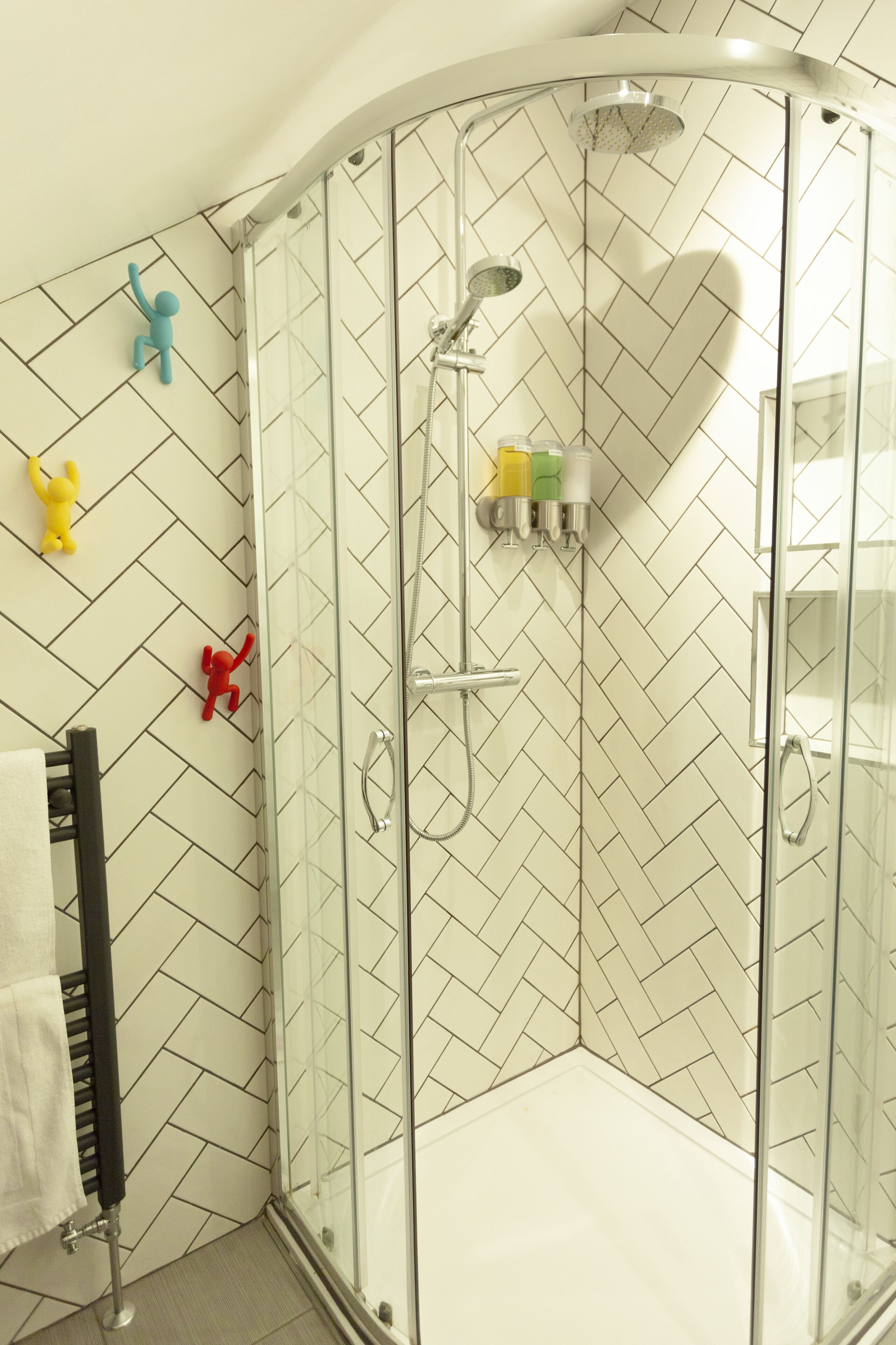 A glass-enclosed shower with a herringbone tile pattern, a towel rack, and a soap dispenser.